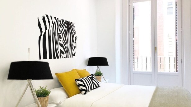 A bedroom with a bed, two bedside tables with lamps, a zebra wall art, and a window with a view of buildings. - Design apartment in Plaza Mayor 3D (Madrid)