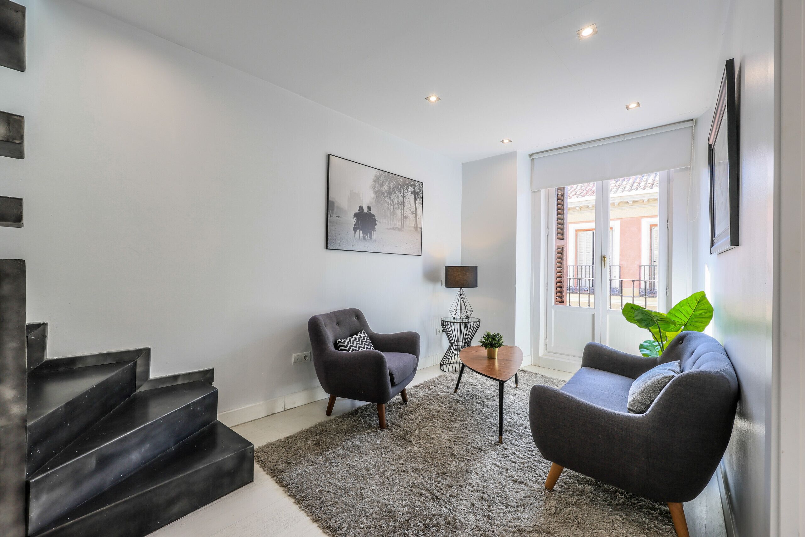 Una sala de estar moderna con una escalera, un sillón gris, un sofá, una mesita y una planta verde grande.
