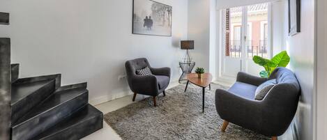 Una sala de estar moderna con una escalera, un sillón gris, un sofá, una mesita y una planta verde grande.