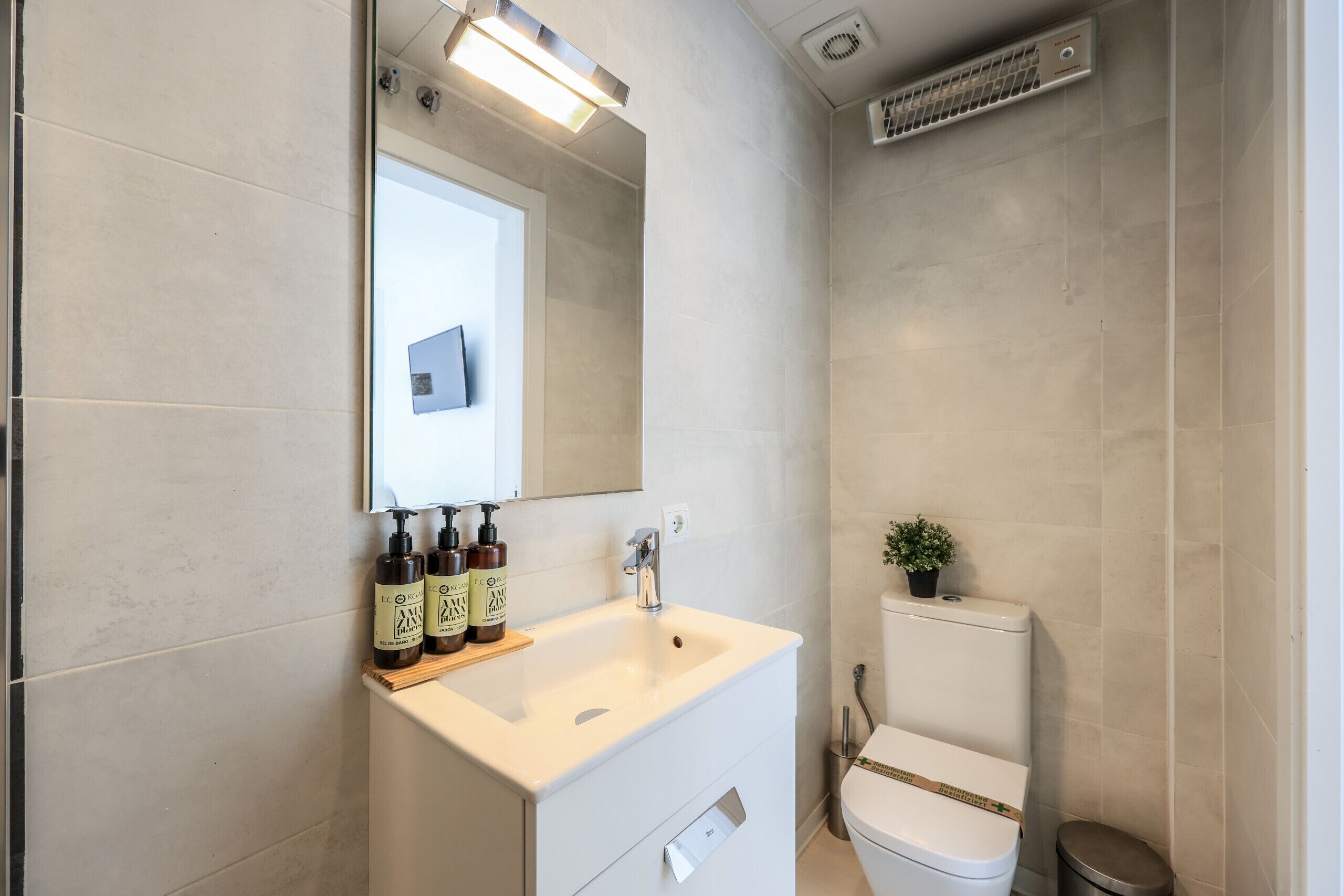 Een moderne badkamer met een wit wastafel, spiegel en toilet. Er staan toiletartikelen op het aanrecht, een klein plantje op het toilet en een ventilatie rooster aan het plafond.
