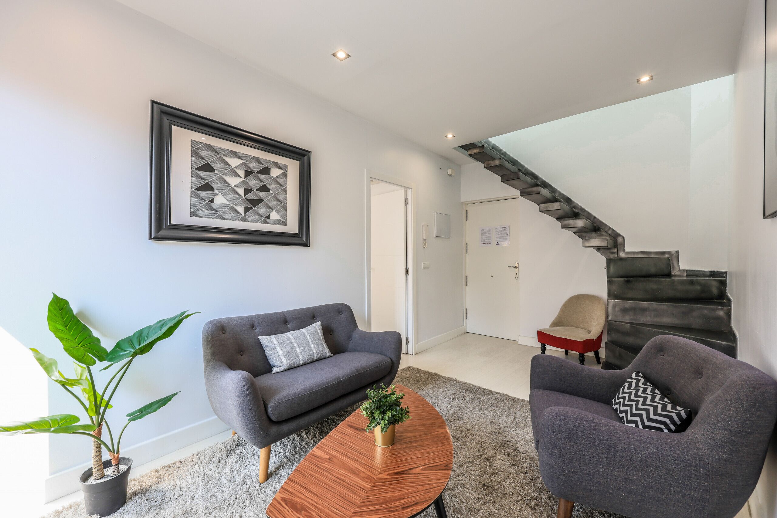 Ein modernes Wohnzimmer mit einem grauen Sofa, einem runden Couchtisch aus Holz und einer Topfpflanze. Im Hintergrund ist eine Treppe mit Metallgeländer zu sehen.
