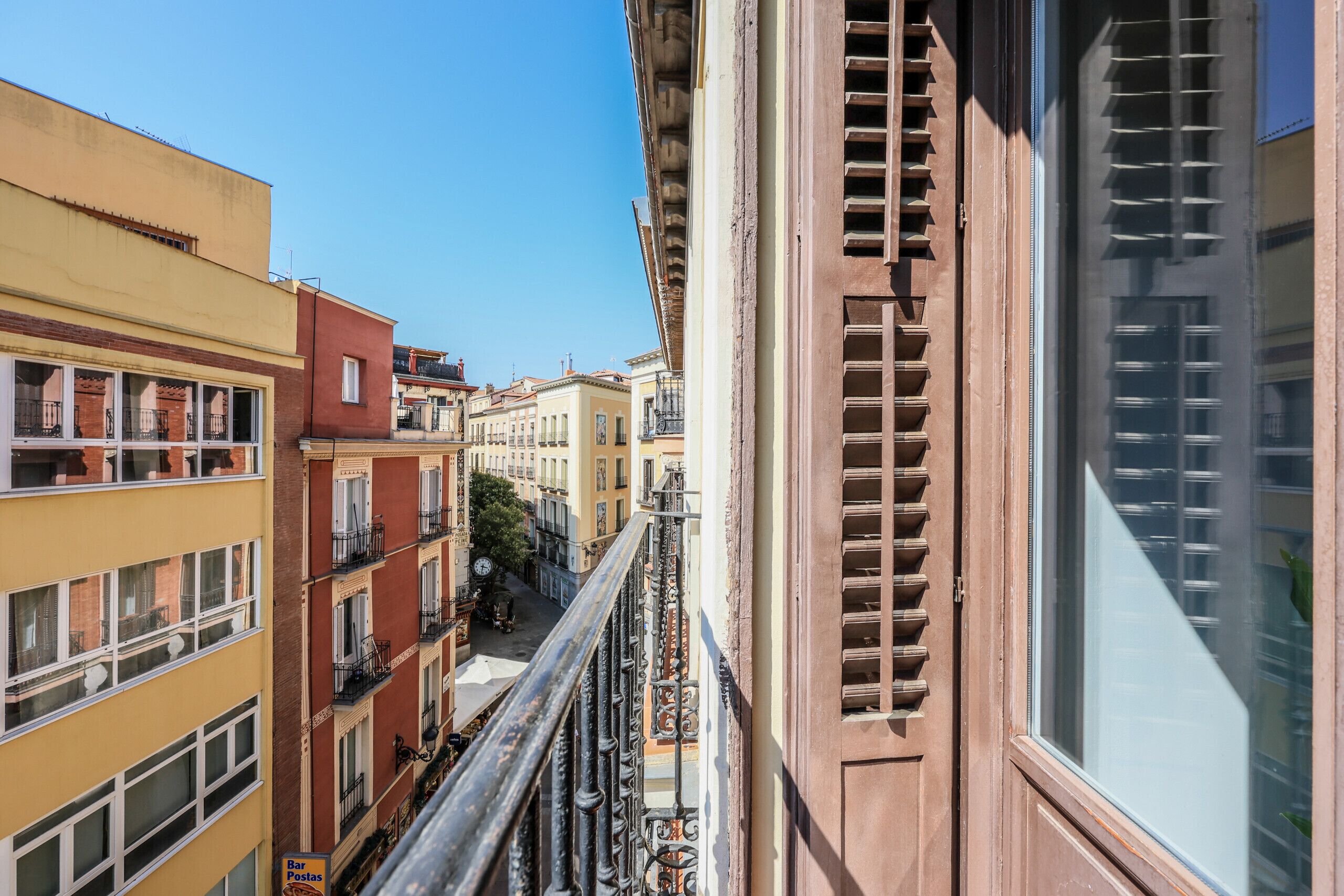 A narrow street with multi-story buildings, balconies, and a clear sky.