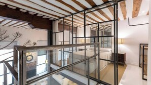 A modern interior with a glass railing overlooking a bedroom, wooden beams on the ceiling, and a visible bed.