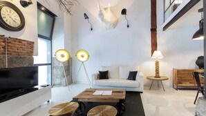 A modern living room with a white sofa, a coffee table, and a television. There are two floor lamps and a wall-mounted light. A large clock and a decorative wall piece are also visible.