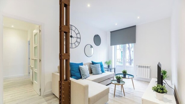 Un salon moderne avec un canapé, une table basse et un téléviseur. Il y a des horloges au mur et une fenêtre donnant sur un beau paysage.