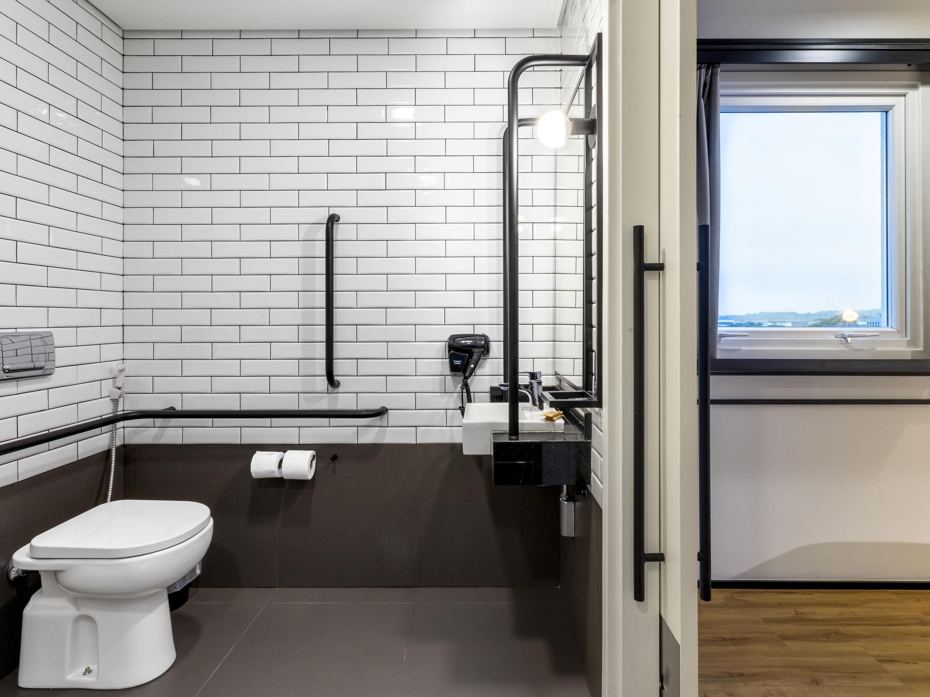 A modern bathroom with white subway tiles, a white toilet, a black grab bar, a towel rack, and a window with a view.