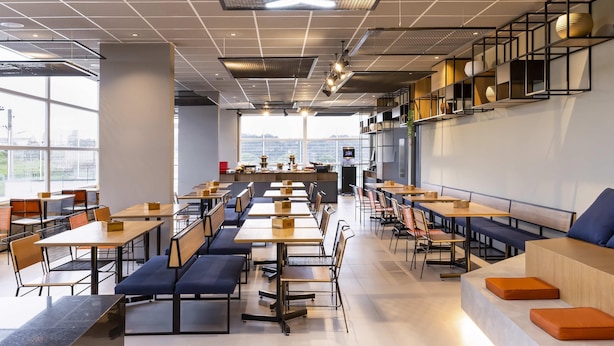 Uma cafeteria moderna com mesas e bancos compridos, um relógio digital e vista para a cidade através de grandes janelas.