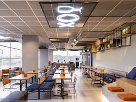 Uma cafeteria moderna com mesas e bancos compridos, um relógio digital e vista para a cidade através de grandes janelas.