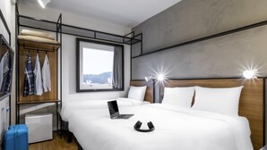 Quarto de hotel com uma cama, um laptop, uma mala e uma janela com vista.