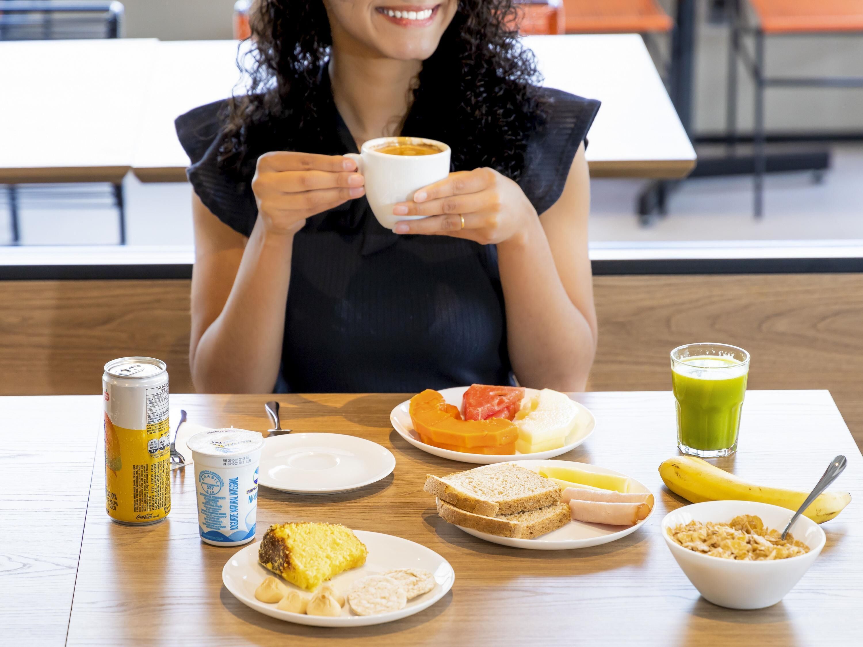 A person sitting at a table with a meal consisting of bread, cheese, ham, a slice of cake, a banana, a glass of juice, a can of soda, and a cup of coffee.