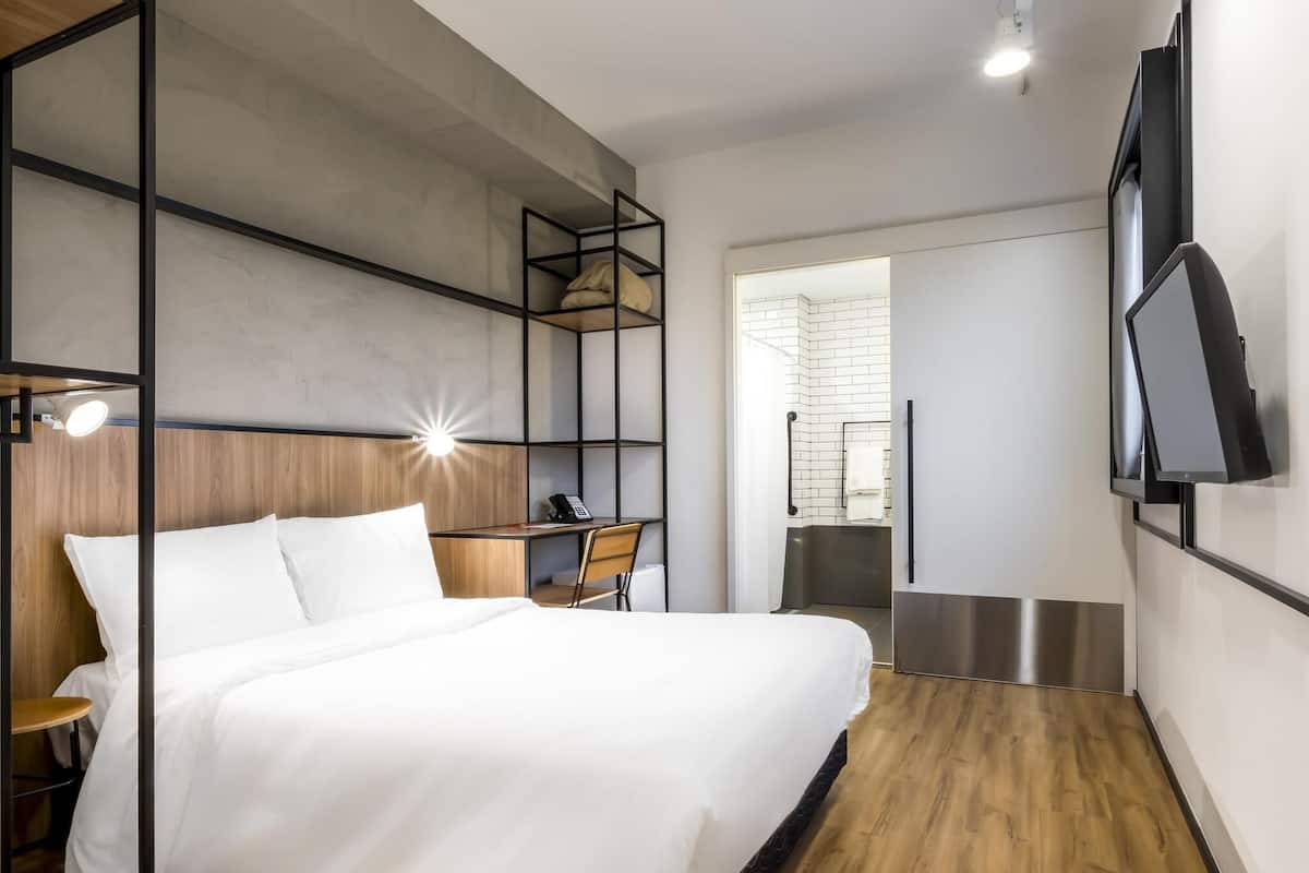 A modern hotel room with a bed, a desk, a chair, a TV, and a shower area.