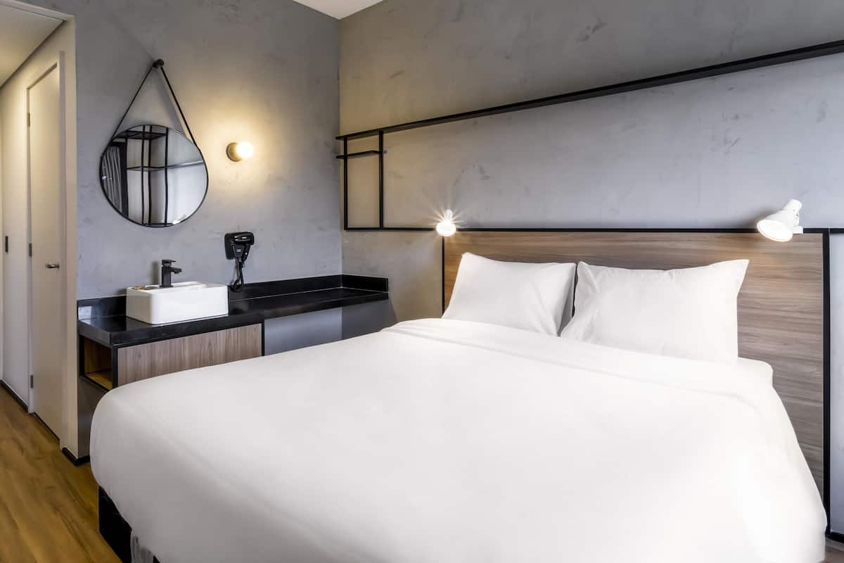 Una habitación de hotel moderna con una cama grande, una mesita de noche con lámpara, un espejo redondo y un estante empotrado.