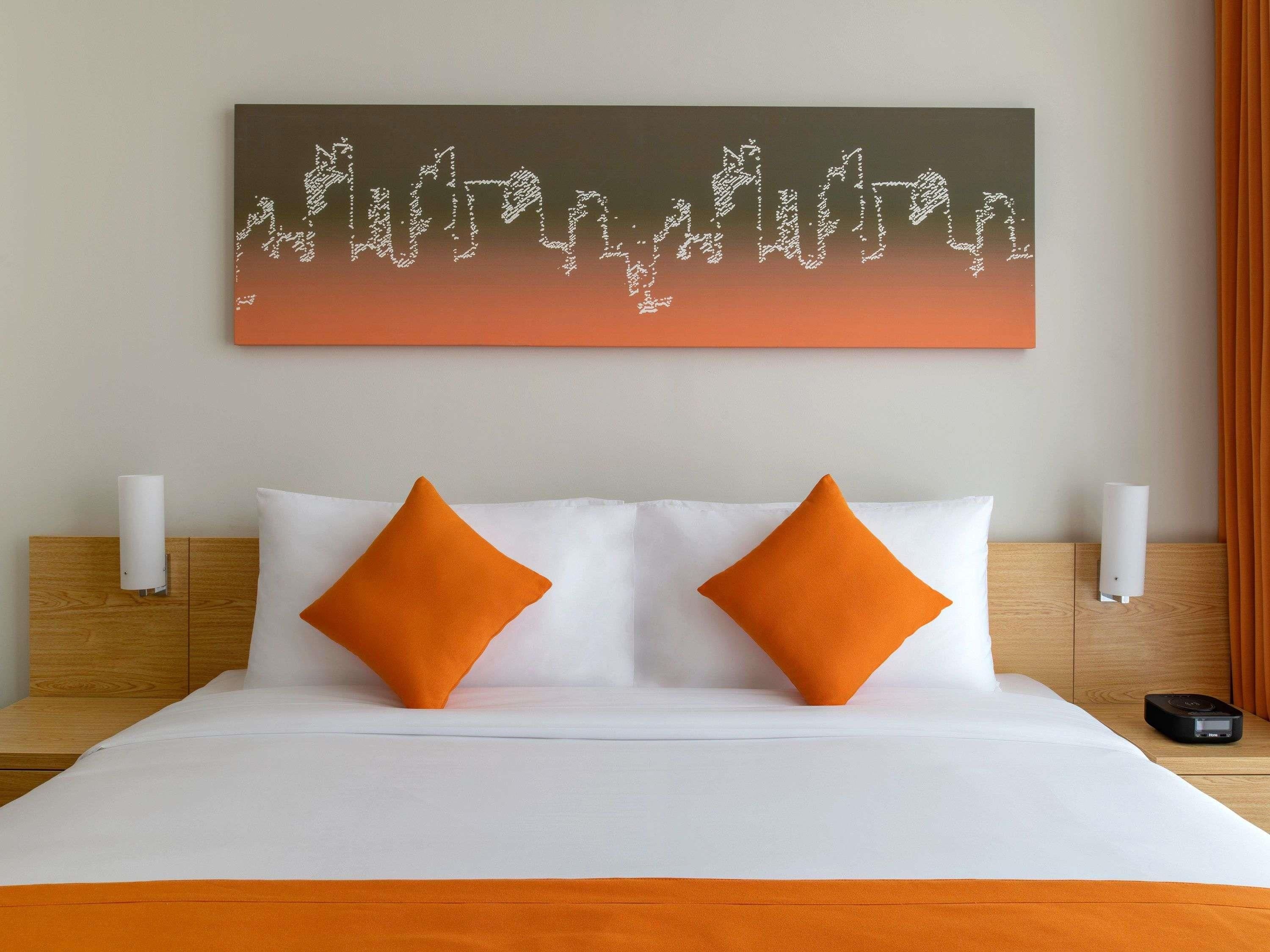 A hotel room with a bed, two pillows, a wall-mounted artwork, and a bedside table with a lamp and a device.