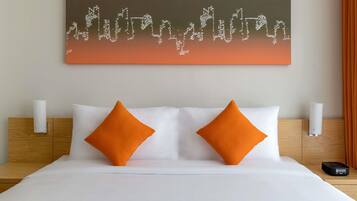 A hotel room with a bed, two pillows, a wall-mounted artwork, and a bedside table with a lamp and a device.