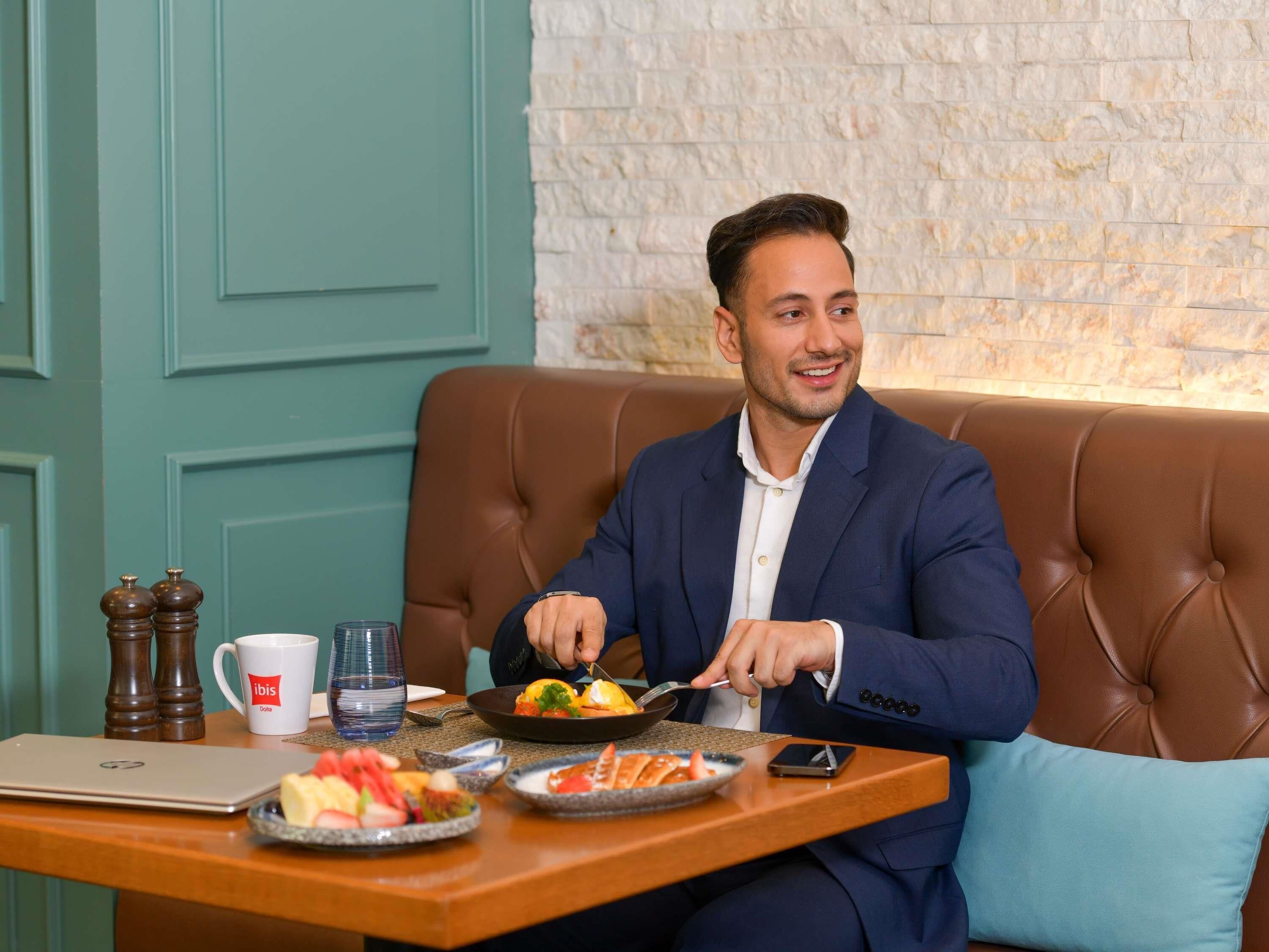 A man sitting on a brown leather couch at a wooden table with a laptop, a glass of water, and plates of food.