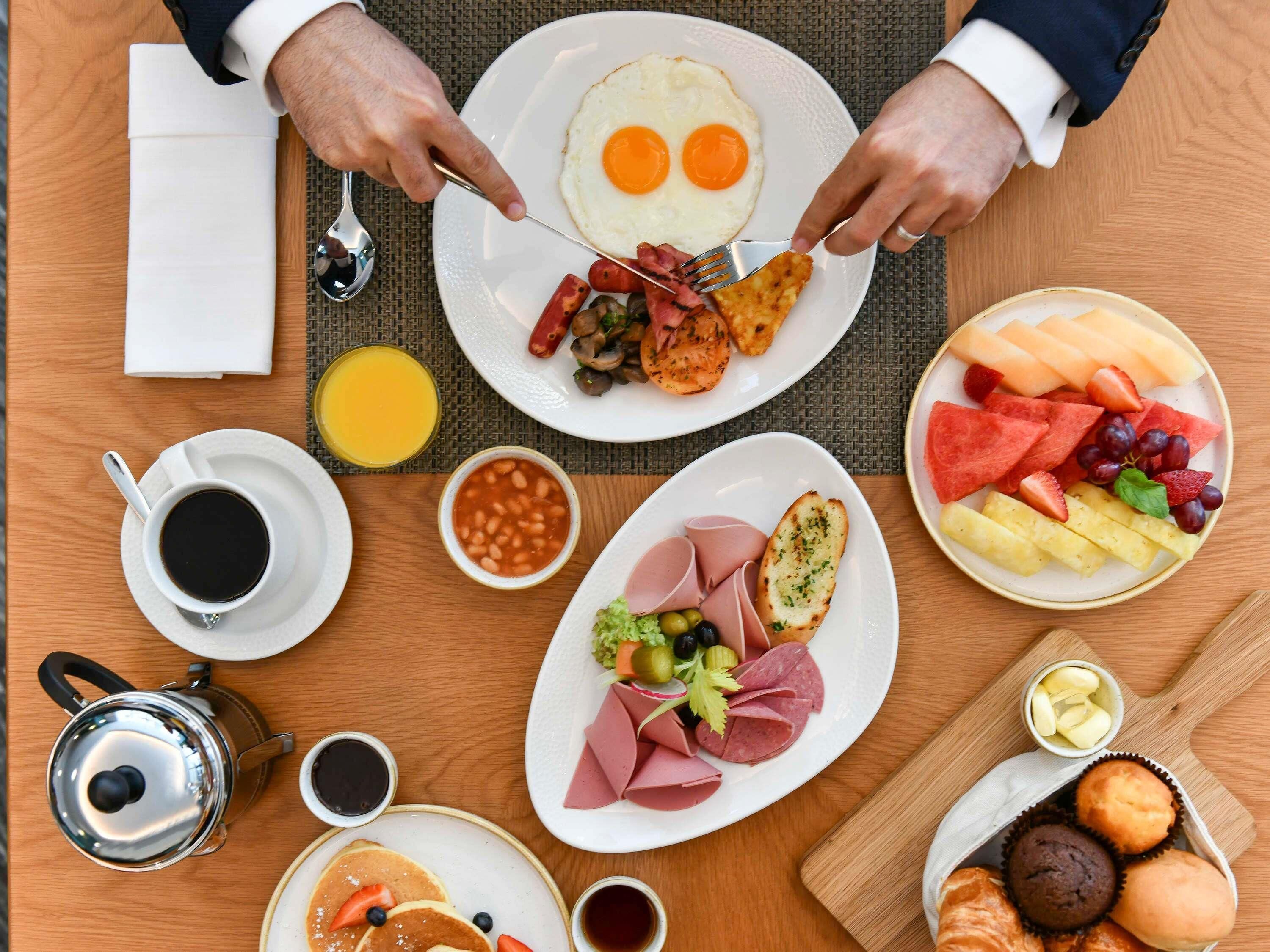 A breakfast setting with plates of eggs, sausages, beans, ham, and various fruits.
