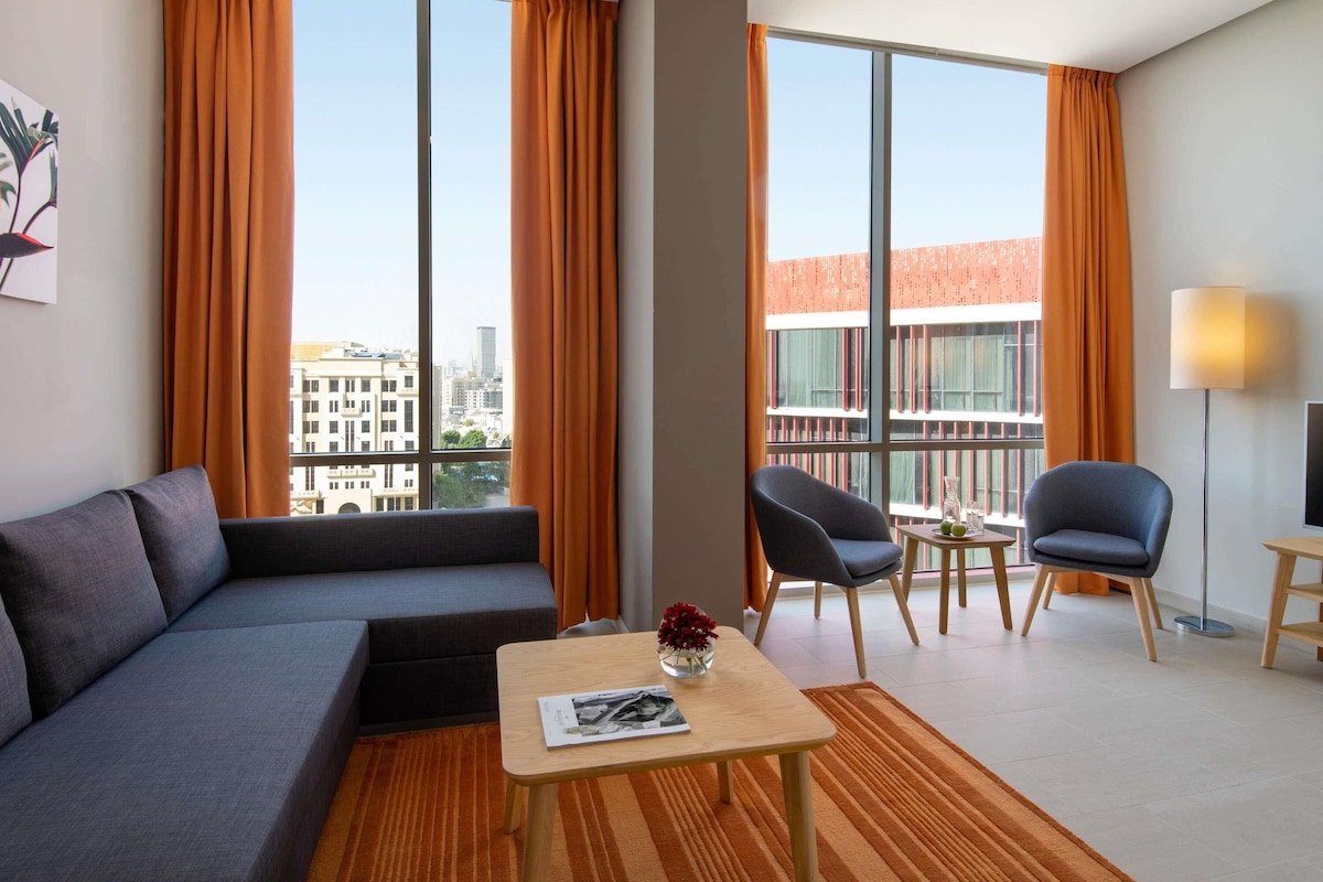 Ett modernt hotellrum med en grå soffa, ett litet träbord, en stol och stora fönster med orangefärgade gardiner.