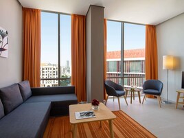 Una habitación de hotel moderna con un sofá gris, una mesita de madera, una silla y ventanas amplias con cortinas anaranjadas.