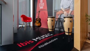A modern interior with a red chair, a guitar, and two drums on a black floor with "ibis Music Doha" branding.