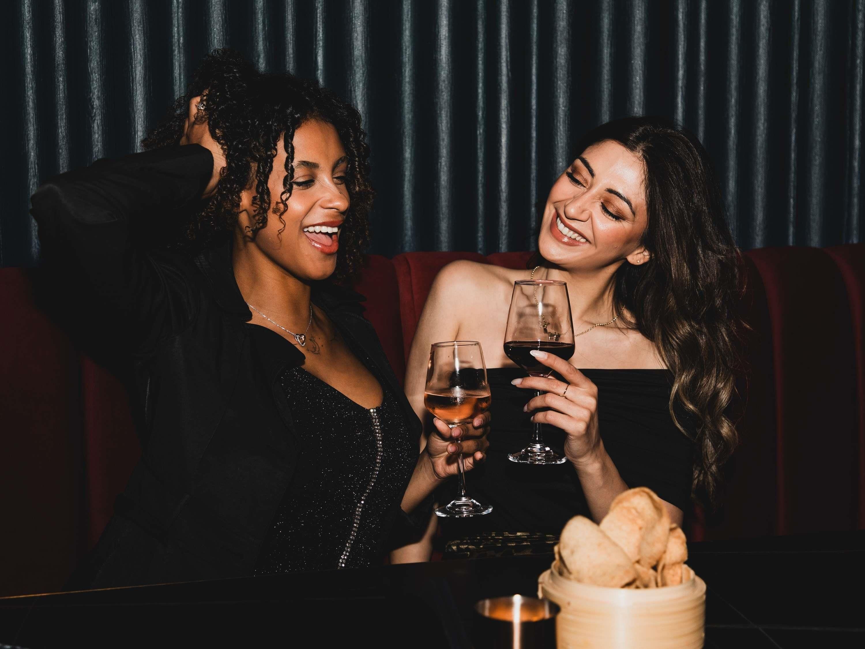 Two people sitting at a table with wine glasses, a basket of bread, and a dark curtain in the background.