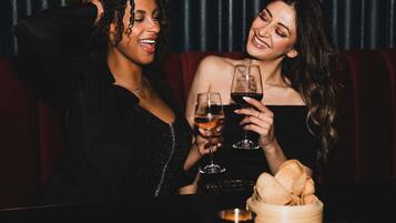 Two people sitting at a table with wine glasses, a basket of bread, and a dark curtain in the background.