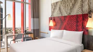 A hotel room with a large bed, a desk with a chair, and a view of the city through large windows.