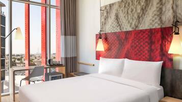 Una habitación de hotel moderna con una cama grande, dos lámparas de noche, un escritorio con silla y vistas a la ciudad a través de amplias ventanas.