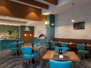 A restaurant with teal chairs and wooden tables, a bar area, and a brick wall with a black and white artwork.