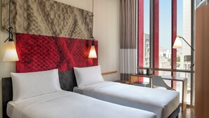 Hotellihuone, jossa on kaksi sänkyä, punainen, kuvioitu seinä, näköala kaupunkiin ja työpöytä, jossa on lamppu.