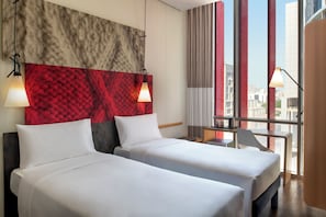 A hotel room with two beds, a red textured wall, a city view, and a desk with a lamp. - Ibis Doha (Doha)