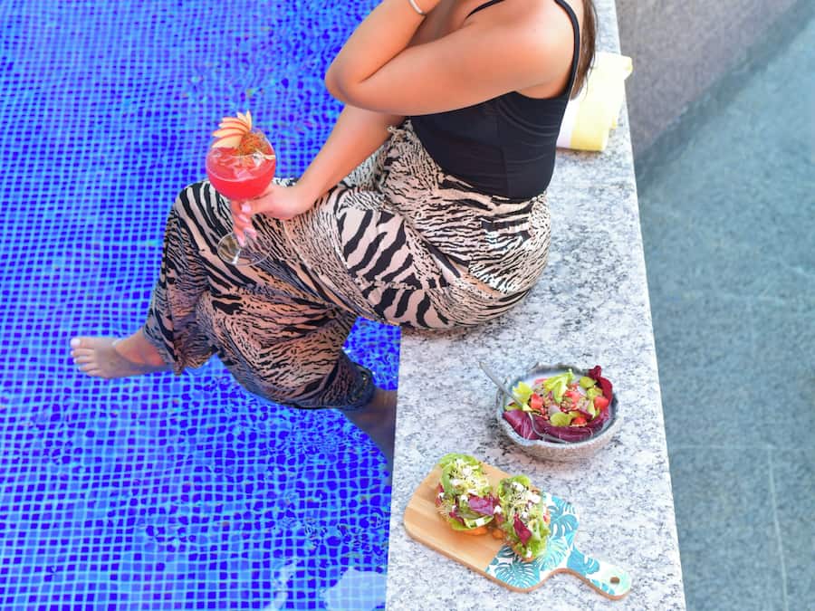 A person sitting by a pool with a drink and a salad on a cutting board.