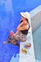 Una persona sentada junto a una piscina, con una bebida y una ensalada sobre una tabla de cortar.