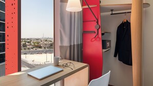 A hotel room with a desk, a laptop, a pair of glasses, a hanging coat, and a view of the city.