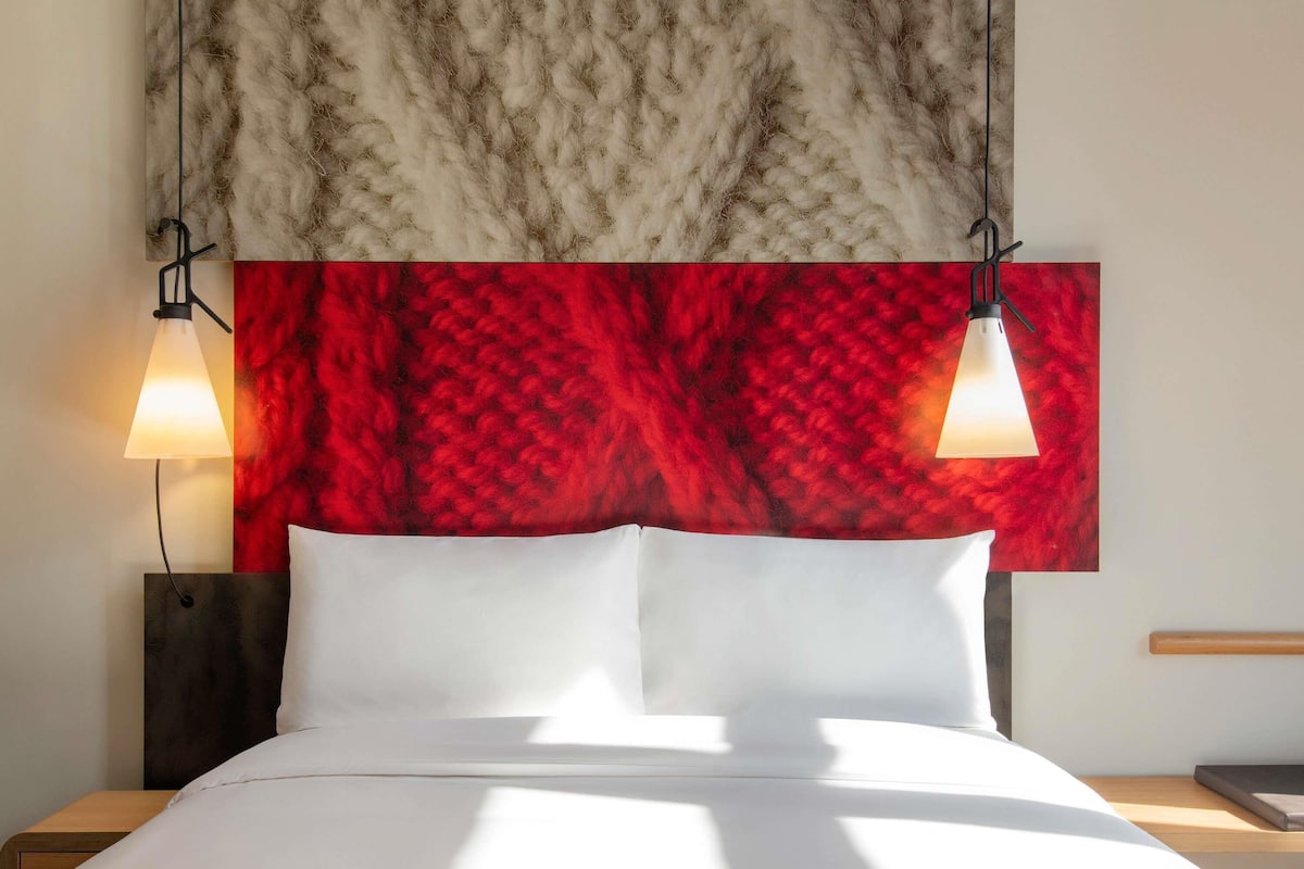 Una habitación de hotel con una cama grande, dos lámparas de noche, un cabecero con un panel texturizado rojo y blanco, y una mesita de noche de madera.
