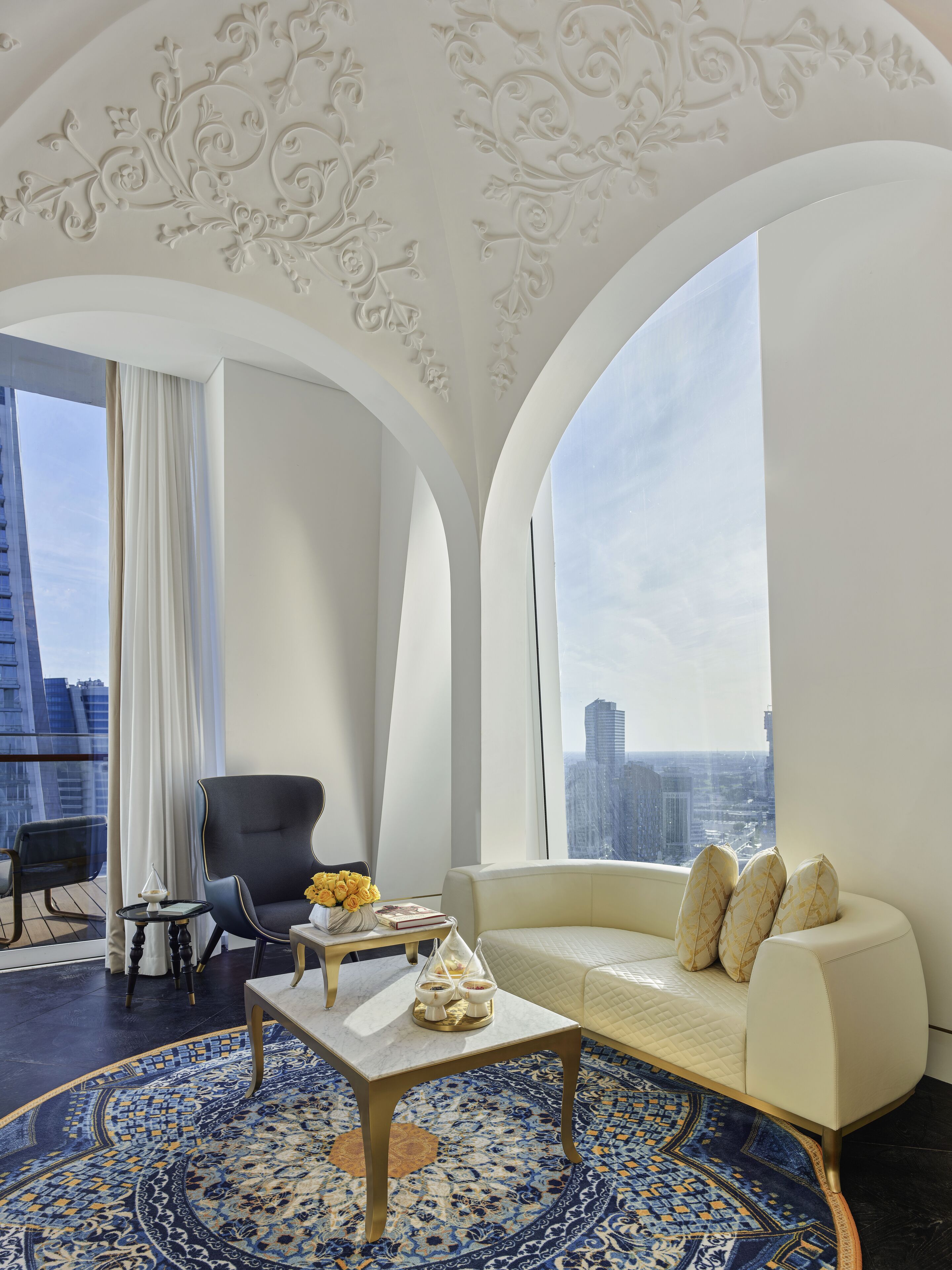 Un salon moderne doté d’un plafond haut, d’un canapé, d’un fauteuil, d’une table basse et offrant une vue sur le paysage urbain grâce à de grandes fenêtres.