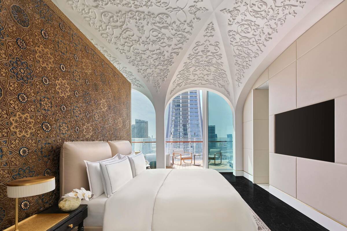 A bedroom with a large bed, a nightstand, a lamp, and a view of a cityscape through a large window.