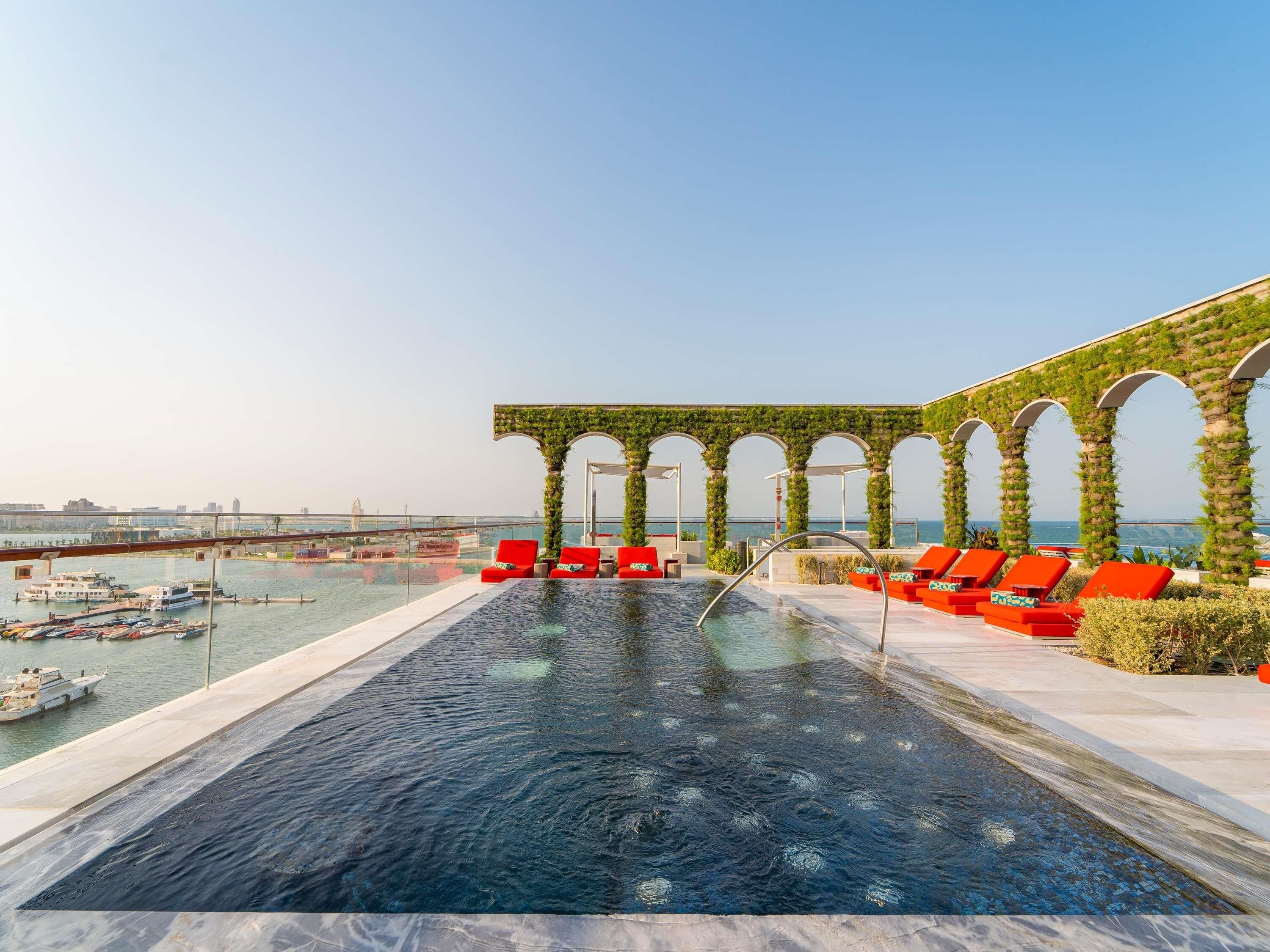 Piscina infinita no terraço com cadeiras de descanso vermelhas e vista para o mar.