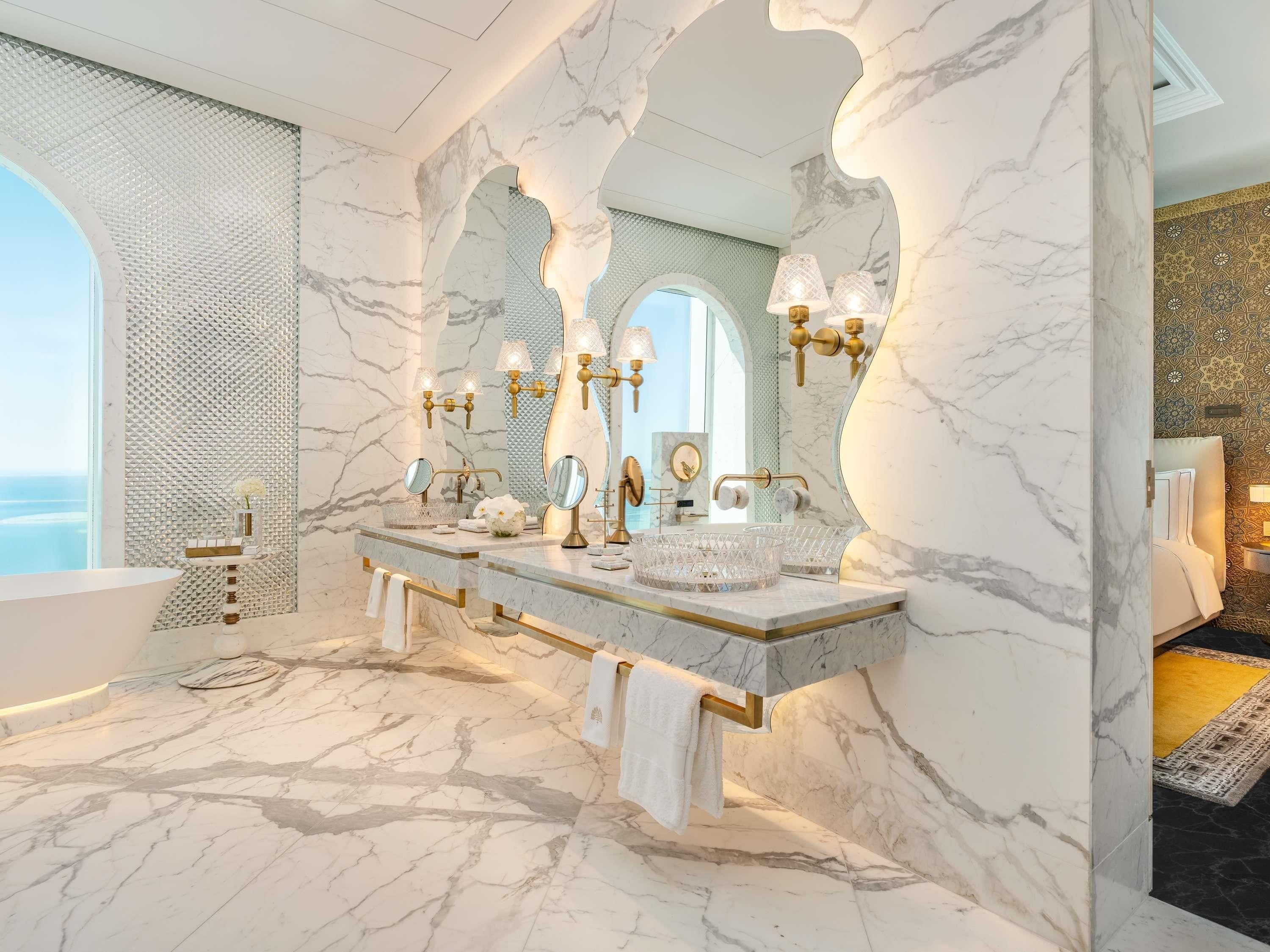 A luxurious bathroom with marble floors, a large vanity with multiple mirrors, and a view of the sea.
