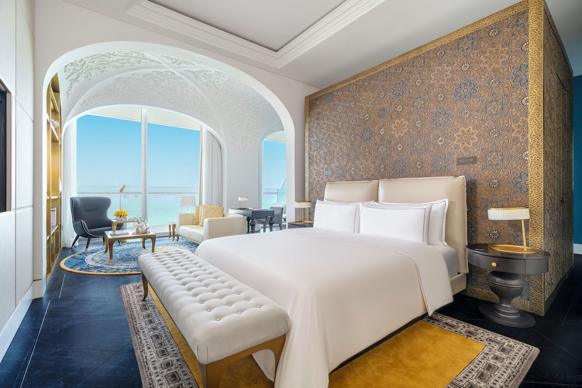 A spacious hotel room with a large bed, a sitting area with a sofa and armchairs, a small table, and a view of the sky through a large window.