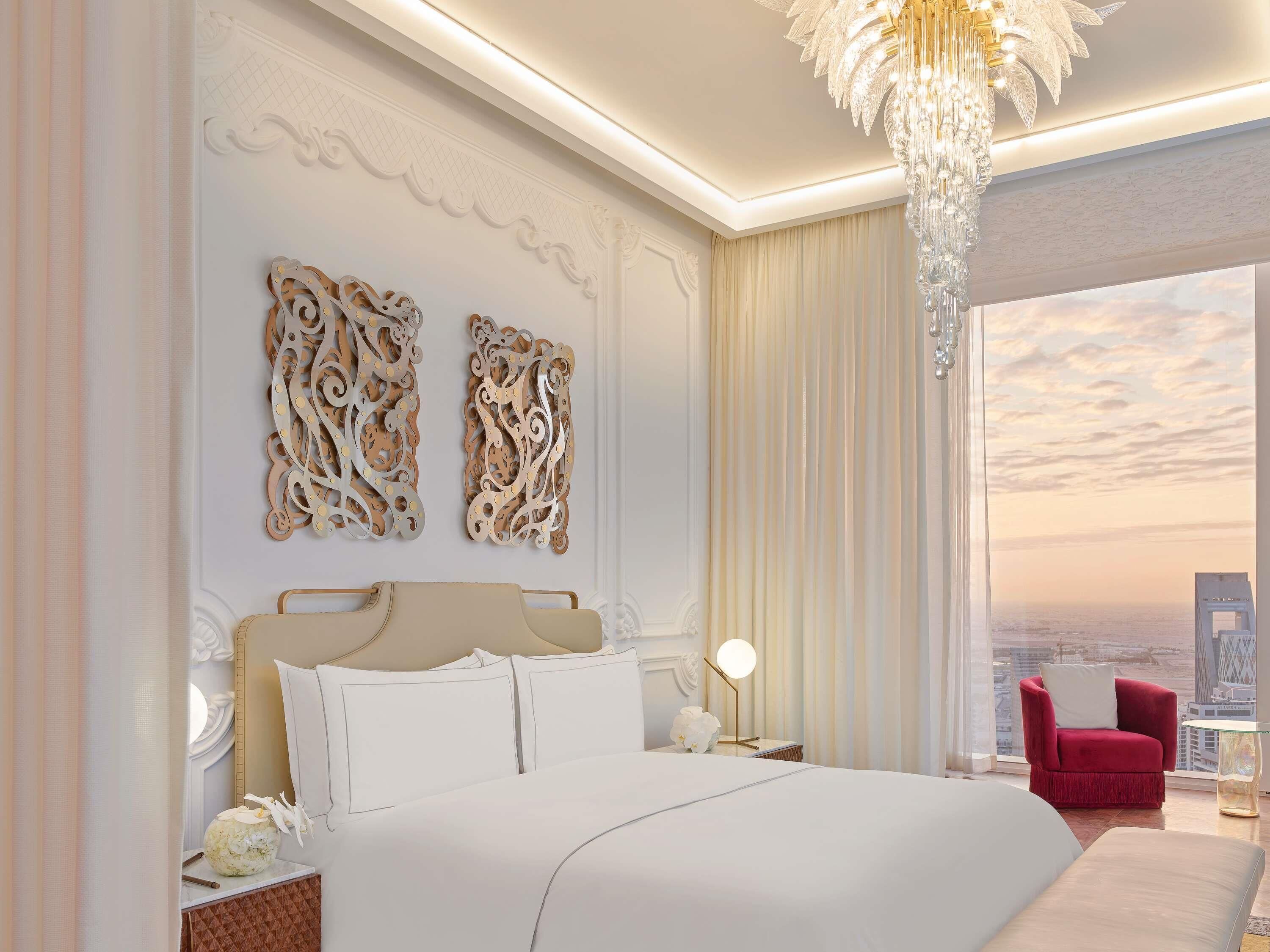 A bedroom with a large bed, decorative wall art, a chandelier, a red chair, and a city view.