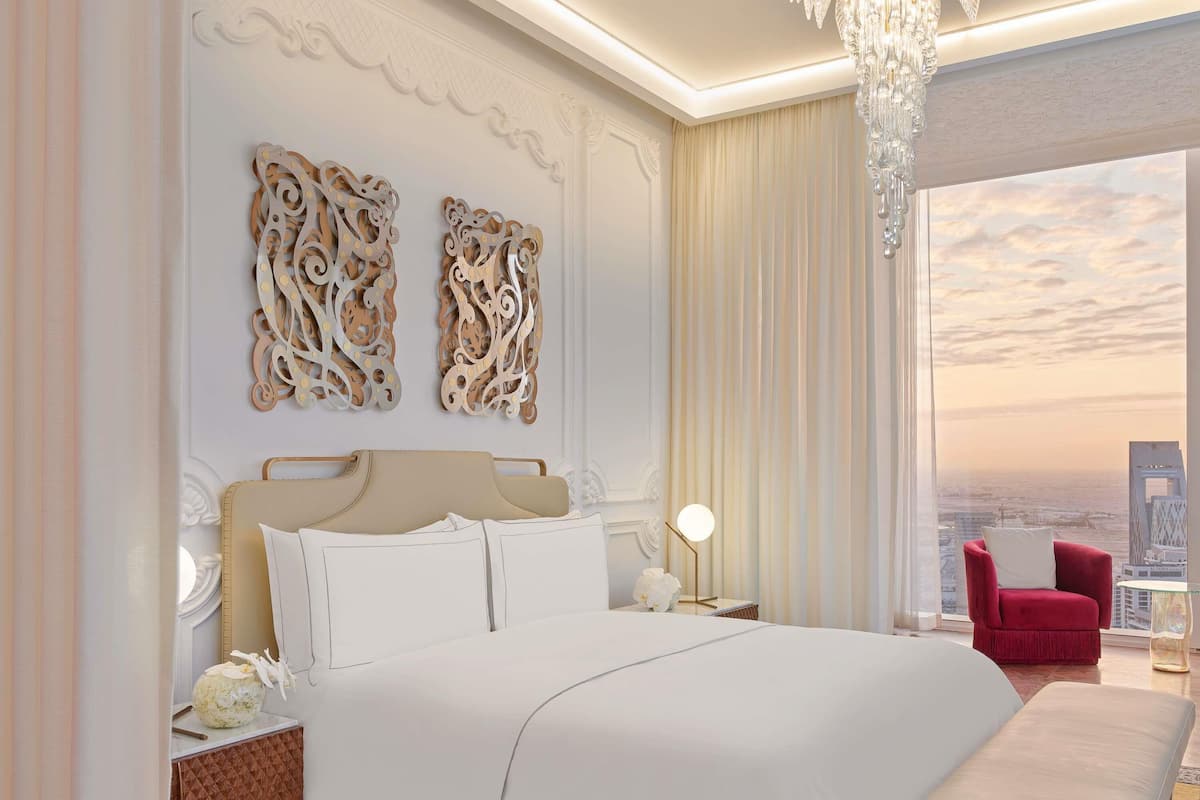 A bedroom with a large bed, decorative wall art, a chandelier, a red chair, and a city view.