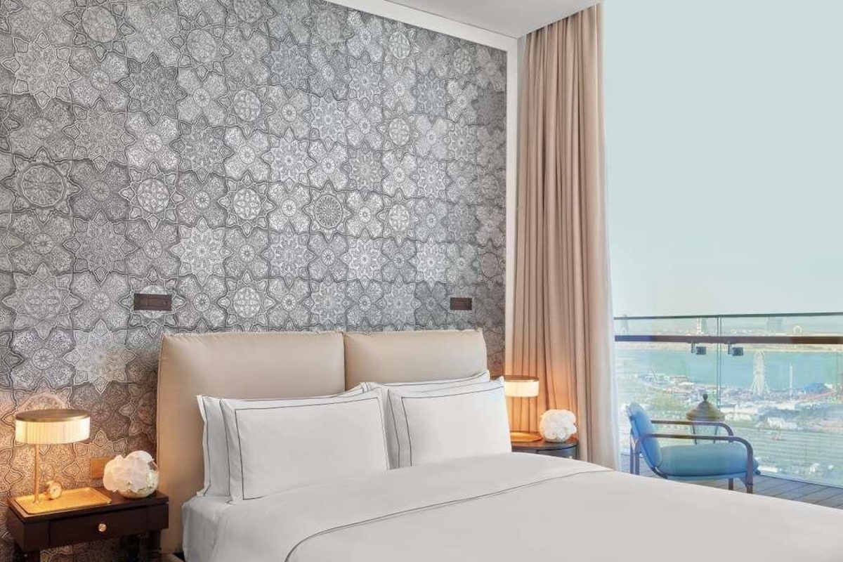 A hotel room with a patterned wallpaper, a bed with white linens, two bedside lamps, a nightstand with a lamp, a blue chair, and a view of a cityscape through large windows.