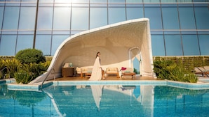 A pool area with a curved white structure, lounge chairs, and a view of a modern building with large windows. - Raffles Doha (Doha)