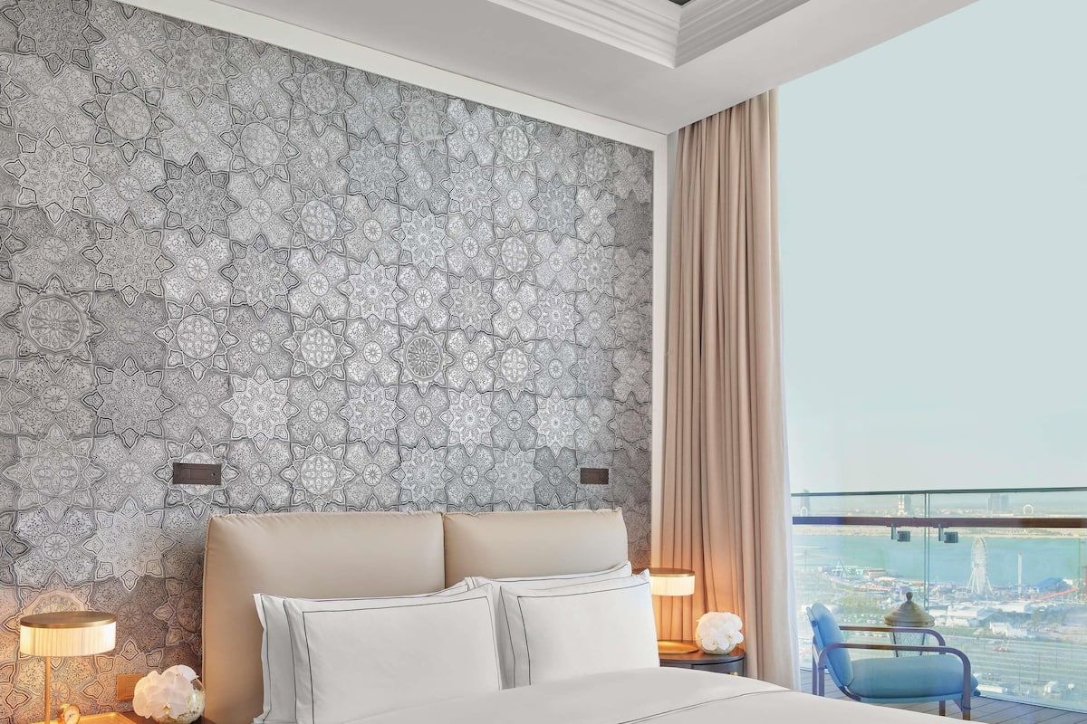 A hotel room with a patterned wallpaper, a bed with white linens, a nightstand with a lamp, and a view of a cityscape through a window.