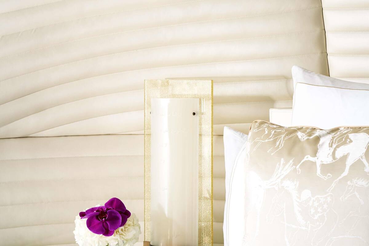 Uma cama com cabeceira branca, uma mesinha de cabeceira com um vaso de flores e um quadro encostado na parede.