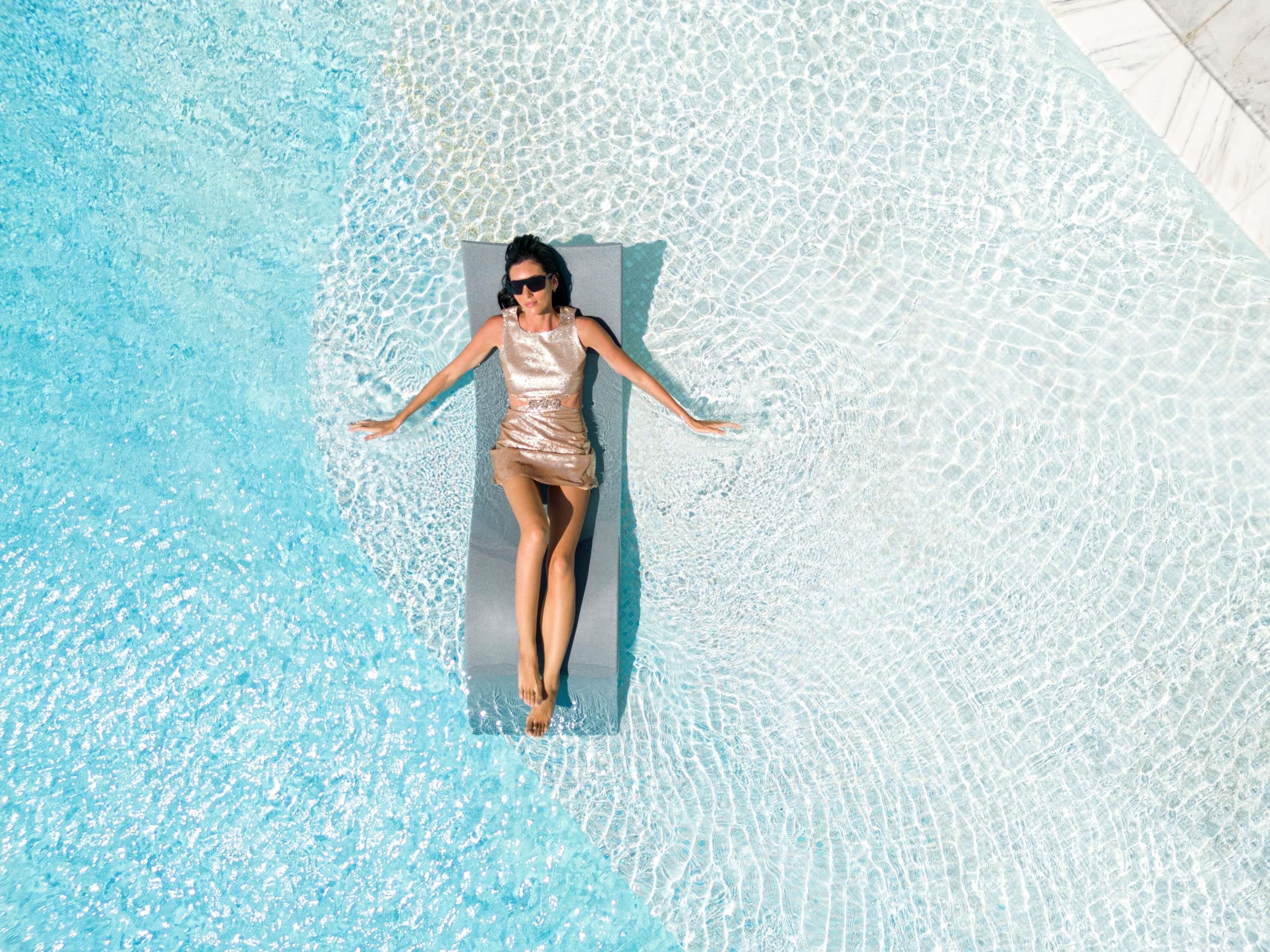 Una persona recostada sobre una plataforma flotante en una piscina de color azul claro.