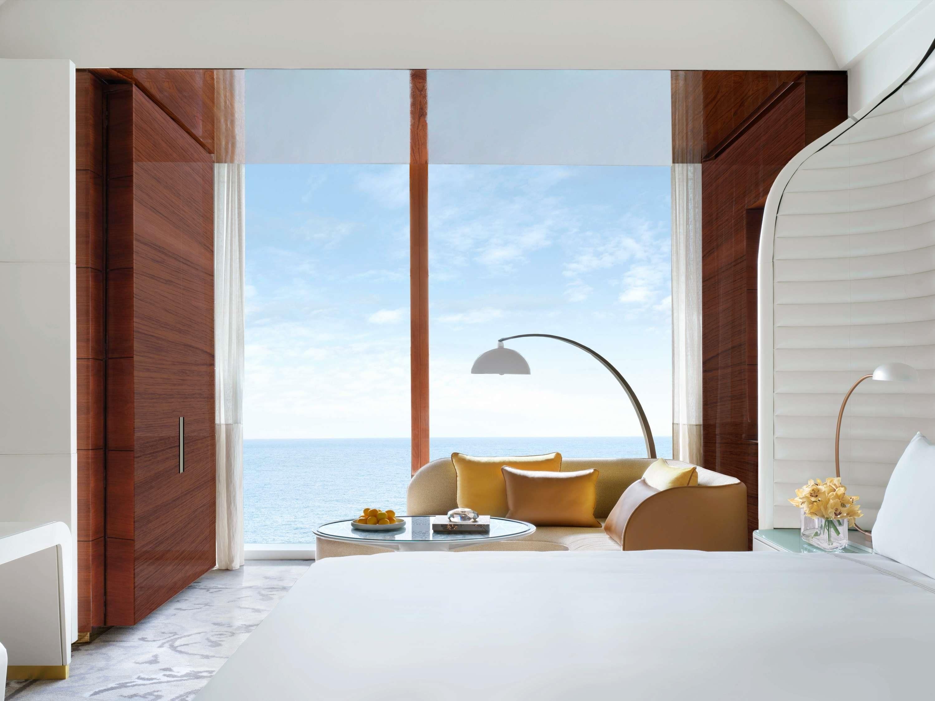 A modern hotel room with a large bed, a sofa, a small table with a lamp, and a view of the sea.