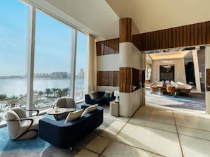Una habitación de hotel moderna con un gran ventanal que ofrece vistas a un cuerpo de agua y al paisaje urbano, una zona de estar con sofás y sillones, y una sala de conferencias con una larga mesa.