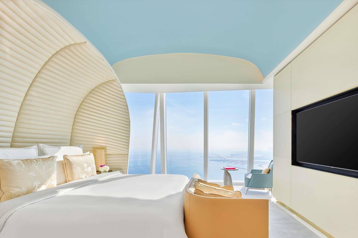 A modern hotel room with a large bed, a seating area, and a large window offering a view of the sea.