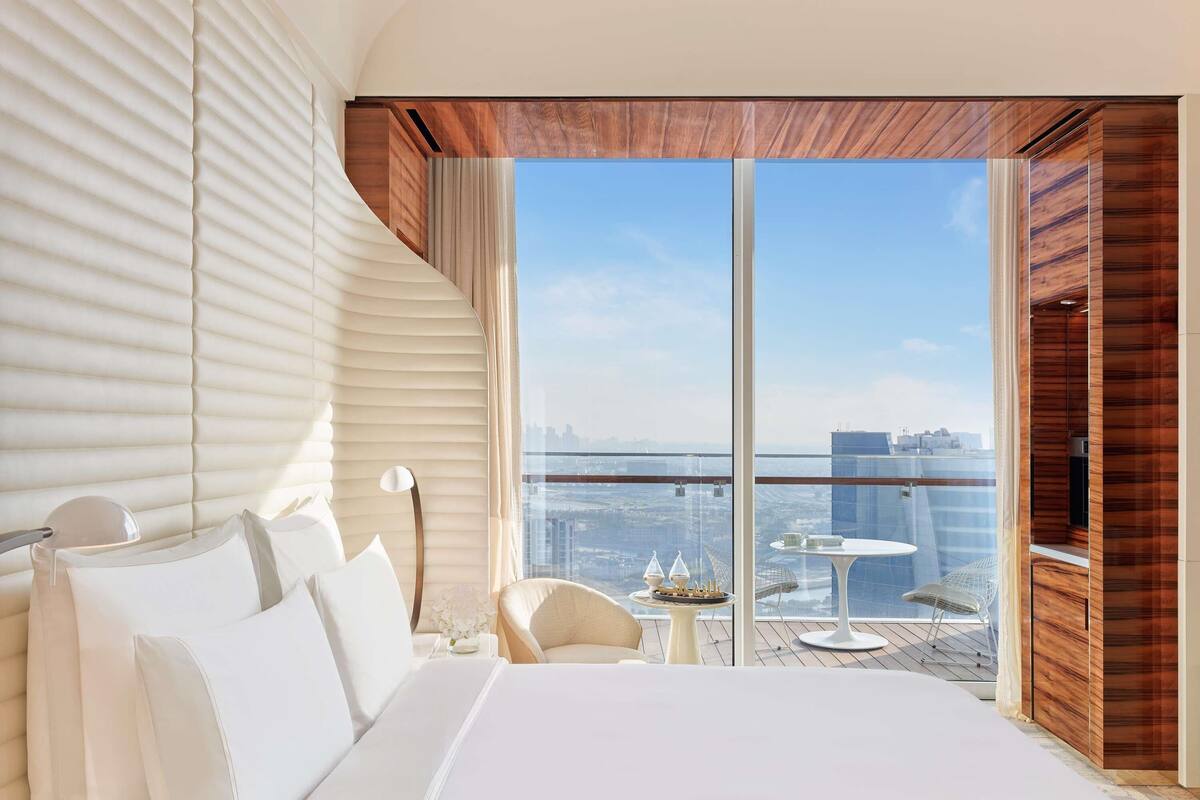 Quarto de hotel moderno com uma cama grande, vista da cidade através de janelas do chão ao teto e uma varanda com mesa e cadeiras.